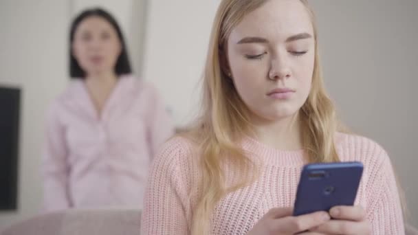 Närbild porträtt av irriterad vit flicka med hjälp av smartphone medan vuxna brunett kvinna skriker på bakgrunden. Mor läskig tonårsdotter hemma. Missförstånd, konflikt. — Stockvideo