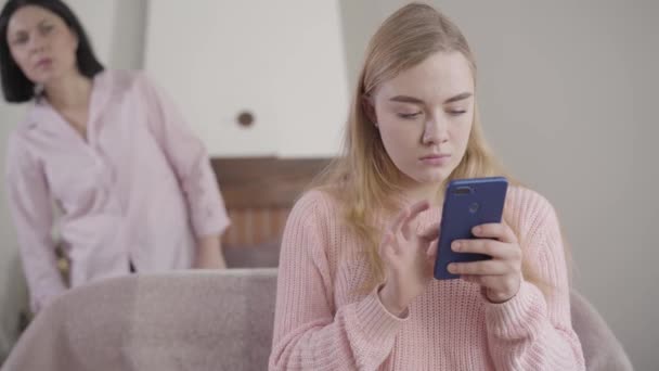 Camera approaching to worried Caucasian girl typing on smartphone as her mother looking at screen from the background. Shocked mom spying teenage daughter. Reliance, relationship. — Stock Video