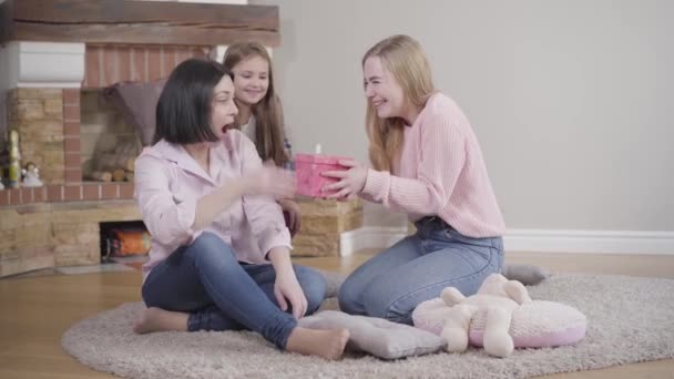 Portrait of happy Caucasian brunette mother receiving gift from daughters. Younger girl opening moms eyes closed with her hands and elder one giving her present. Cheerful family resting at home. — Stock Video