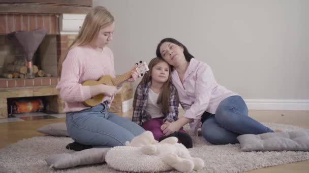 Csinos szőke kaukázusi lány ukulelét játszik otthon anyának és a húgának. Tehetséges tinédzser, aki hétvégenként szórakoztatja a családot. Életmód, szabadidő, hobbi, — Stock videók