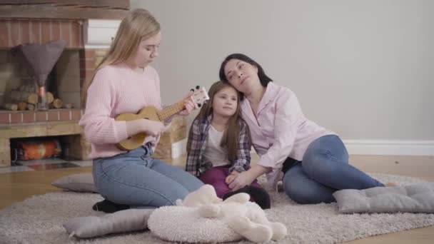 Mooi blond blank meisje dat ukelele speelt voor moeder en jonge zus thuis. Getalenteerde tiener onderhoudend gezin in het weekend. Lifestyle, vrije tijd, hobby, — Stockvideo