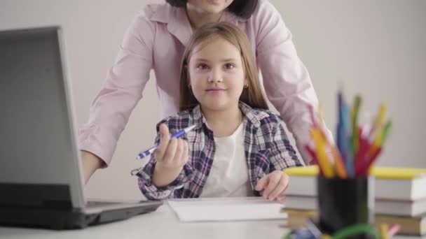 Kameran närmar sig en vacker vit skolflicka som sitter vid bordet och håller pennan. Brunett oigenkännlig mor står på rygg. Stöd, utbildning, studier. — Stockvideo