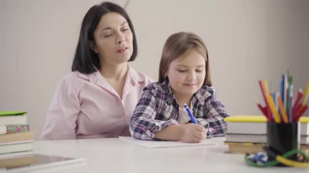 Retrato de una linda colegiala caucásica sentada con mamá y escribiendo en un libro de ejercicios. Madre morena ayudando a su hija con la tarea. Apoyo, educación, estudio . — Vídeo de stock