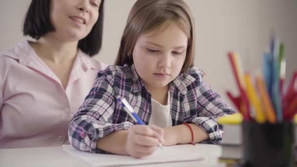 Közelkép egy csinos barna, fehér iskolás lányról, aki anyával ül és a füzetébe ír. Felnőtt mosolygós anya segít a lányának a házi feladatban. Támogatás, oktatás, tanulás. — Stock videók