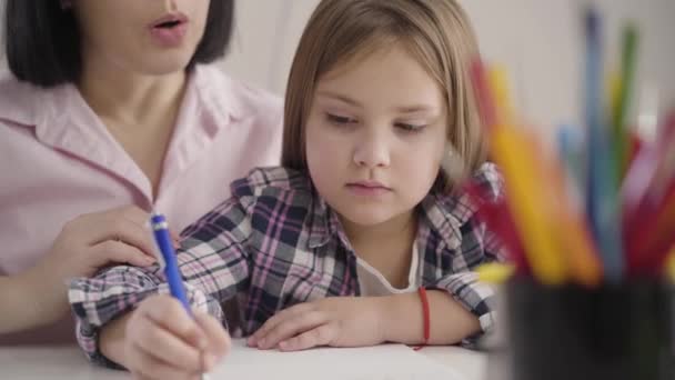 Närbild av koncentrerad kaukasisk brunett flicka skriver i träningsboken som hennes oigenkännliga mamma dikterar i bakgrunden. Skolflicka gör läxor med mamma inomhus. — Stockvideo