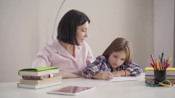 若い白人の母親は娘にかなりブルネットの女の子が書き留めるように指示しています。子供は家で親と宿題をしている。支援、教育、知性. — ストック動画