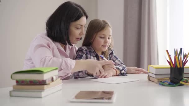 Side view van brunette Kaukasische moeder helpen dochter op te schrijven. Mooi schoolmeisje doet huiswerk met mama thuis. Ondersteuning, onderwijs, intelligentie. — Stockvideo