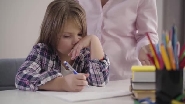 Zbliżenie portretu zmęczonej białej dziewczyny siedzącej przy stole i piszącej. Nierozpoznawalna kobieta ściskająca dłonie w tle. Mama beszta córkę za problemy w edukacji. — Wideo stockowe