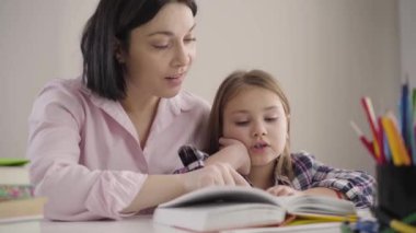 Annesiyle birlikte evde kitap okuyan beyaz bir kızın yakın çekimi. Mutlu okul kızı ailesiyle ödev yapıyor. Anne kızının çalışmasına yardım ediyor. Zeka, eğitim, destek.