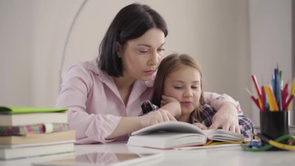 Portrait de fille caucasienne intelligente livre de lecture pour maman. Mignon écolier faisant ses devoirs avec sa mère. Un parent qui aide sa fille à étudier. Intelligence, éducation, soutien . — Video