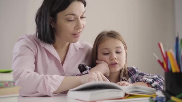 Zbliżenie uroczej białej dziewczyny czytającej książkę z mamą w domu. Szczęśliwa uczennica odrabiająca lekcje z rodzicami. Matka pomaga córce w nauce. Inteligencja, edukacja, wsparcie. — Wideo stockowe