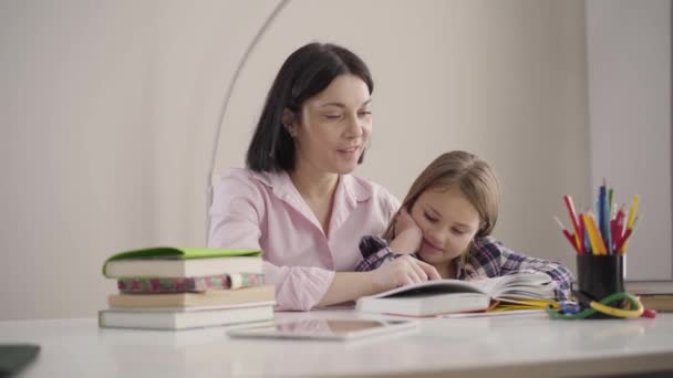 Porträtt av lycklig vit mor läsa bok för dotter och kyssa hennes huvud. Positiv skolflicka som gör läxor med hjälp av föräldrar. Utbildning, intelligens, hobby. — Stockvideo