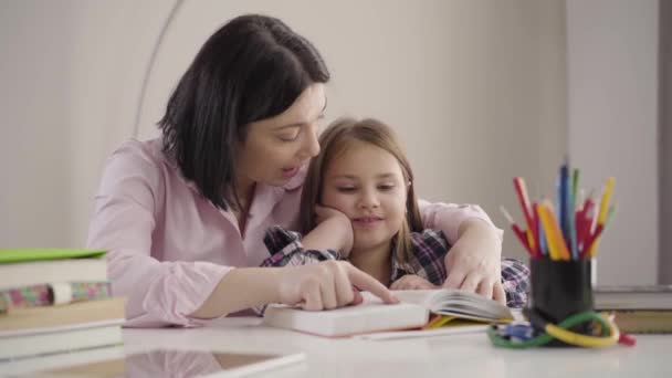 Kameran närmar sig vit mor och dotter läser tillsammans. Skolflicka läser bok med mamma hemma. Mor hjälper dottern att studera. Intelligens, utbildning, stöd. — Stockvideo