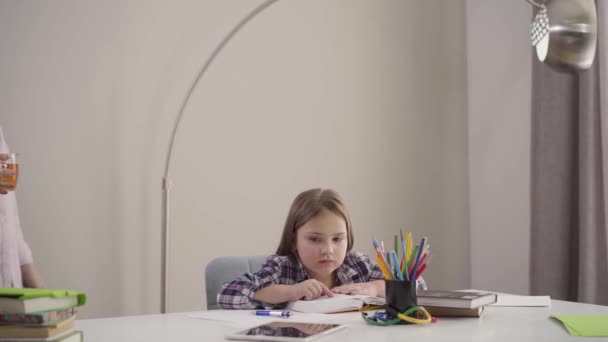 En söt vit tjej som sitter vid bordet och läser som mamma och kommer med en kopp te. Föräldrarna tar hand om dottern. Utbildning, stöd, intelligens. — Stockvideo