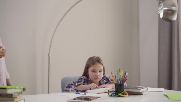 Chica bastante caucásica sentada en la mesa y leyendo como mamá viene con una taza de té. Padres cuidando de su hija. Educación, apoyo, inteligencia . — Vídeos de Stock