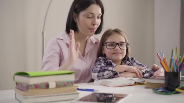 Porträtt av glada kaukasiska mor och dotter ger high five, tittar på kameran och ler. Glad skolflicka i glasögon och vuxen kvinna sitter vid bordet med läxor. — Stockvideo