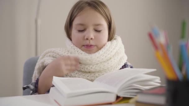 Nahaufnahme Porträt eines charmanten kaukasischen Mädchens, das mit Buch im weißen Schal sitzt und niest. kranke Schülerin beim Hausaufgabenmachen. Bildung, Überlastung, Gesundheitsversorgung. — Stockvideo