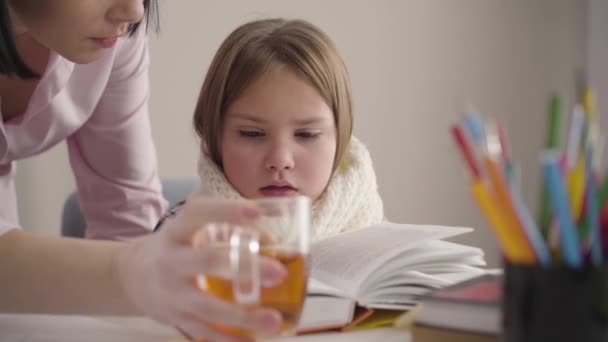 Знімальний портрет хворої кавказької школярки, що сидить за столом і читає, як мама йде з чашкою чаю. Жінка з Брюнета піклується про дочку. Освіта, підтримка, інтелект. — стокове відео