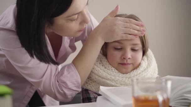 Detailní záběr brunetky bělošské matky kontrolující teplotu dcer. Hodná školačka, co sedí u stolu a čte si. Žena, která se stará o dceru. Vzdělávání, podpora, inteligence. — Stock video