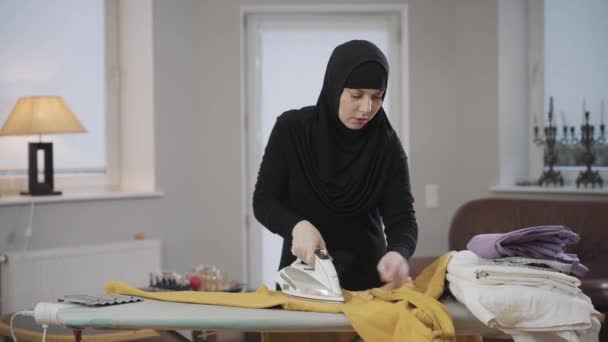 Retrato de mujer musulmana concentrada en hiyab planchando jersey amarillo. Ama de casa haciendo tareas domésticas en interiores. Sociedad patriarcal, cultura oriental . — Vídeo de stock