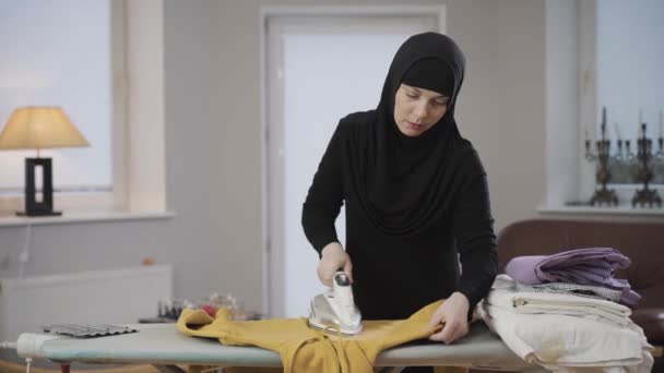 Diligente ama de casa musulmana en hiyab negro planchando suéter amarillo en casa. Joven mujer hermosa haciendo tareas domésticas. Derecho comunitario, derecho comunitario, derecho comunitario . — Vídeos de Stock