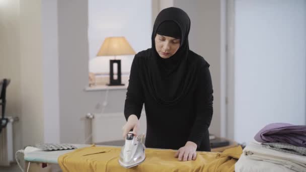 Porträtt av trött muslimsk kvinna i svarta traditionella kläder gör hushållsarbete. Ung vacker dam i hijab strykning och torka pannan med handen. Österländsk kultur, traditioner. — Stockvideo