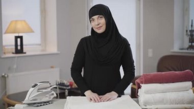 Kameraya bakıp gülümseyen çekici Müslüman kadının portresi. Çamaşırlarla ütü masasının önünde duran siyah tesettürlü ev kadını. Gelenekler, doğu kültürü..