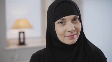 Siyah tesettürlü güzel Müslüman kadının yakın plan portresi kameraya bakıyor ve gülümsüyor. Kahverengi gözlü çekici bayan evde poz veriyor. Kamera soldan sağa hareket ediyor.