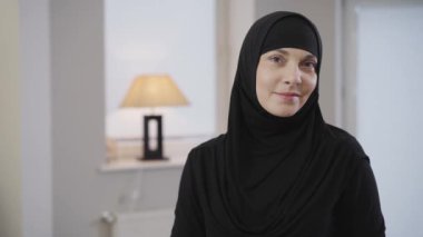 Geleneksel siyah tesettürlü güzel Müslüman kadın kameraya bakıyor ve gülümsüyor. Kahverengi gözlü güzel bir kadının portresi. Doğulu insanlar, gelenekler. Kamera sağdan sola hareket ediyor.