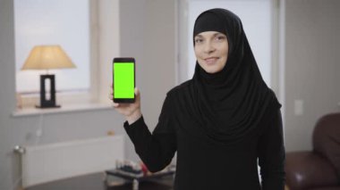 Elinde yeşil ekranla tesettürlü gülümseyen Müslüman kadının portresi. Siyah elbiseli modern doğulu kadın içeride poz veriyor. Motivasyon, doğu kültürü.