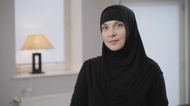 Μέση φωτογραφία της όμορφης Μουσουλμάνας με τη μαύρη μαντίλα να κοιτάζει την κάμερα και να χαμογελάει. Χαριτωμένη κυρία με καστανά μάτια που ποζάρει στο σπίτι με παραδοσιακό φόρεμα. Κάμερα που κινείται από αριστερά προς τα δεξιά. — Αρχείο Βίντεο