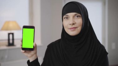 Siyah tesettürlü kendine güvenen Müslüman bir kadının elinde yeşil ekran ile akıllı telefon tutuşu. Geleneksel siyah elbiseli modern güzel bayan içeride poz veriyor. Motivasyonlu, doğulu insanlar.