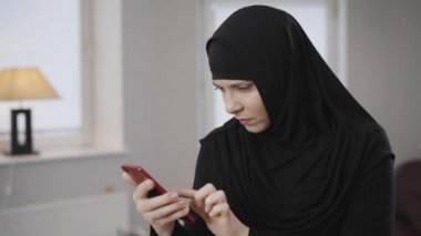 Geleneksel tesettürlü, akıllı telefon ekranına bakıp gülümseyen modern, güzel Müslüman kadın. Evde sosyal medyayı kullanan pozitif kadın. Toplum, yaşam tarzı, doğu kültürü.