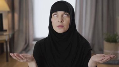 Güzel Müslüman bir kadının yakın plan portresi. Elleri havada oturuyor, yukarı bakıyor ve konuşuyor. Geleneksel tesettürlü genç bayan evde dua ediyor. Din, geleneksel kültür..