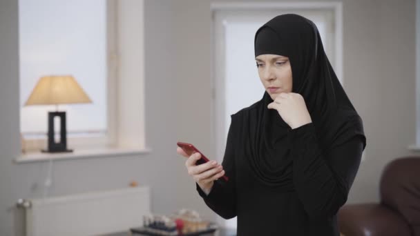 Mujer musulmana seria en hijab mirando la pantalla del teléfono inteligente y desplazándose. Joven dama oriental en vestido tradicional usando el teléfono móvil en casa. Gente oriental, cultura moderna . — Vídeo de stock