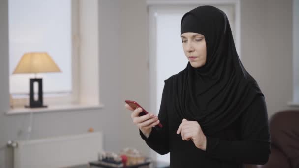 Side view van jonge moslimvrouw met behulp van sociale media op haar smartphone. Lachende moderne dame in hijab die thuis rust. Samenleving, oosterse cultuur, levensstijl. — Stockvideo