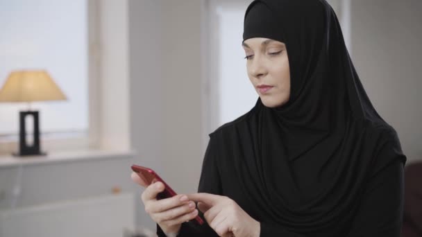 Widok z boku portret młodej muzułmanki przewijającej się na smartfonie i gestykulującej nie trzęsąc głową. Nowoczesna wschodnia kobieta zszokowana mediami społecznościowymi. Kultura tradycyjna, społeczeństwo. — Wideo stockowe