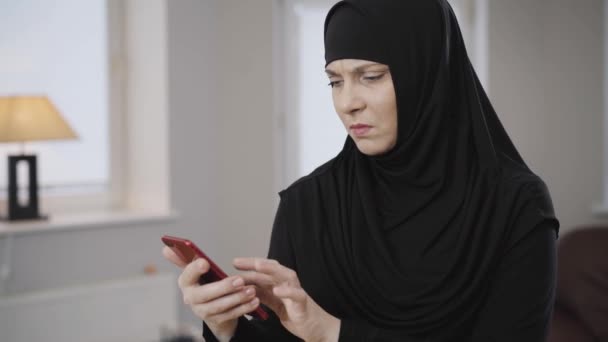 Giovane donna musulmana in hijab tradizionale guardando lo schermo dello smartphone e facendo espressione facciale scioccata. Signora diligente orientale che scuote la testa. Cultura tradizionale, società moderna . — Video Stock