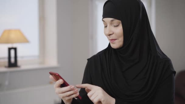 Portrét vzrušené muslimky v tradičním oblečení rolovat na smartphonu a usmívat se. Veselá moderní východní dáma používající sociální média. Společnost, životní styl, východní kultura. — Stock video