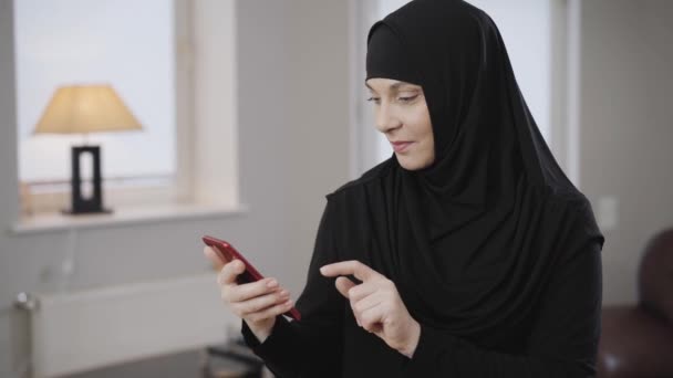 Mujer musulmana moderna de confianza en el hiyab negro tradicional que envía un mensaje en el teléfono inteligente. Señora oriental satisfecha desplazándose en el teléfono inteligente y sonriendo. Sociedad moderna, estilo de vida, cultura oriental . — Vídeo de stock