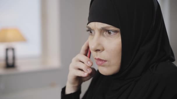 Close-up rosto de mulher muçulmana com olhos castanhos falando no smartphone. Senhora irritada em hijab preto brigando com alguém. Cultura Oriental, problemas . — Vídeo de Stock