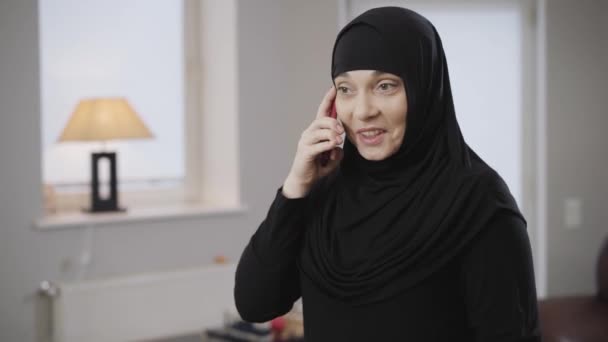 穿着头巾的年轻现代穆斯林妇女在智能手机上说话和在情感上做手势的画像。 东方女士在家里用手机。 现代技术、传统文化. — 图库视频影像