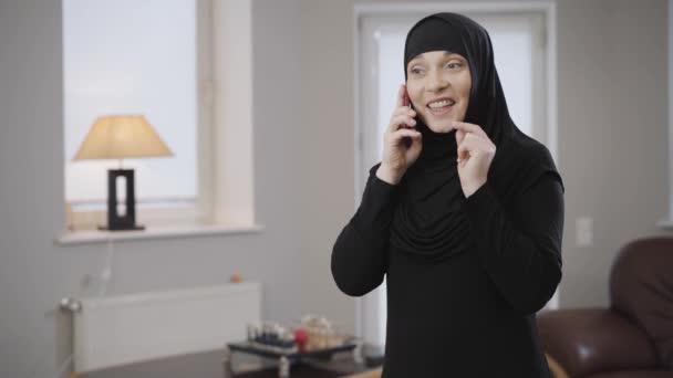 镜头缓缓接近身穿头巾的年轻穆斯林妇女，她用智能手机说话，面带微笑。 现代东方女士使用手机和手势。 现代技术、传统文化. — 图库视频影像