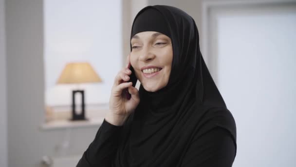 Nahaufnahme einer jungen schönen muslimischen Frau im schwarzen Hijab, die ihr Smartphone zu Hause benutzt. Ost-Dame spricht und lächelt. Moderne Technologien, traditionelle Kultur. — Stockvideo