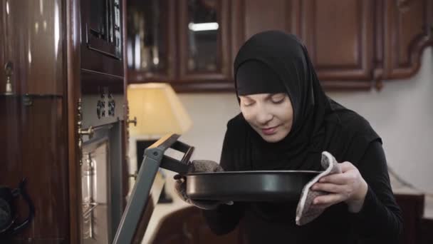 Retrato de una sonriente mujer musulmana vestida de negro tradicional oliendo a comida horneada. Moderna dama oriental en hijab cocinar para la familia en casa. Cultura tradicional, hobby, estilo de vida . — Vídeo de stock