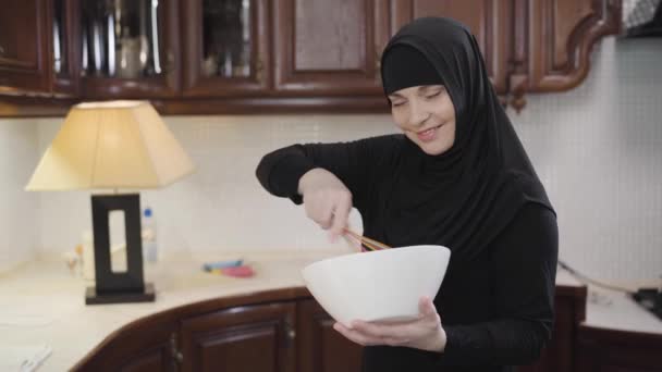 Tiro médio da bela mulher muçulmana no tradicional hijab olhando para a câmera e batendo. Senhora oriental moderna cozinha na cozinha. Sociedade moderna, tradições orientais . — Vídeo de Stock