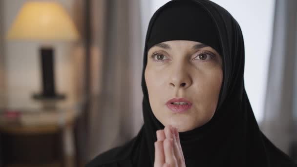 Retrato de cerca de una atractiva mujer musulmana cogida de la mano y rezando. Dama de negro hiyab tradicional hablando con Dios y cerrando los ojos. Cámara moviéndose de izquierda a derecha . — Vídeo de stock