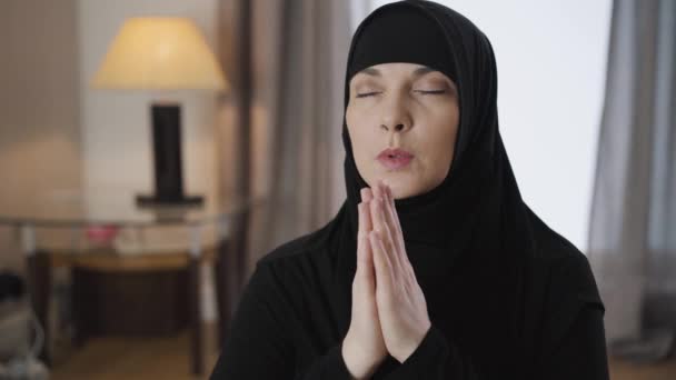 Portrait of young beautiful Muslim woman sitting with closed eyes and holding hands together. Adult lady in black hijab praying at home. Lifestyle, religion. Camera moving around from right to left. — 비디오