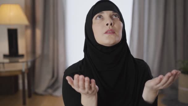 Jonge moslimdame in hijab zittend met opgeheven handen, omhoog kijkend en hoofd schuddend. Oosterse vrouw in zwarte hijab biddend thuis. Religie, traditionele cultuur. — Stockvideo