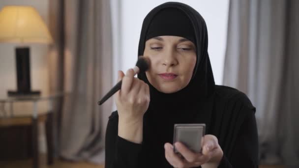 Primer plano de la hermosa mujer musulmana aplicando polvo facial. Linda señora oriental en hiyab negro haciendo maquillaje en casa. Cultura moderna, belleza, cuidado de la cara . — Vídeo de stock